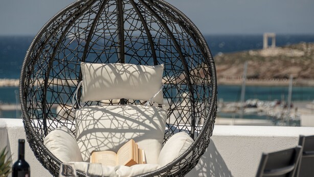Un fauteuil suspendu en osier, doté de coussins blancs et accompagné d’un livre, offre une vue imprenable sur la côte, avec des bateaux et un bâtiment situé au loin.
