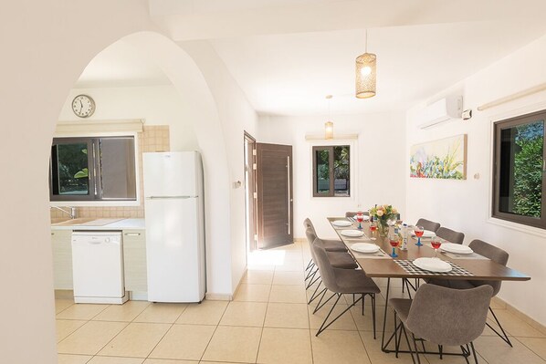 Une cuisine moderne avec des appareils électroménagers blancs, un coin repas avec une longue table et des chaises, et une vue sur la verdure par une fenêtre.