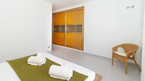 Ein Hotelzimmer mit einem Bett, einer grünen Matte, zwei gerollten Handtüchern, einem Korbstuhl und einem Holzschrank.