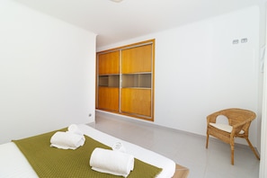 A hotel room with a bed, a green mat, two rolled towels, a wicker chair, and a wooden wardrobe.
