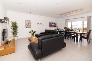 Una sala de estar moderna con un sofá negro, una mesa de centro de madera y un comedor con una mesa y sillas negras. Hay plantas en macetas y obras de arte en las paredes.