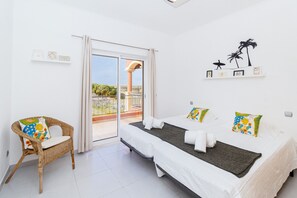 Ein Schlafzimmer mit zwei Betten, einem Korbstuhl, einem Balkon mit Blick und einer Wand mit gerahmten Bildern.