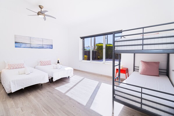 Una habitación con dos camas individuales, una litera, un ventilador de techo, una ventana con vistas y una mesita de noche con lámpara.