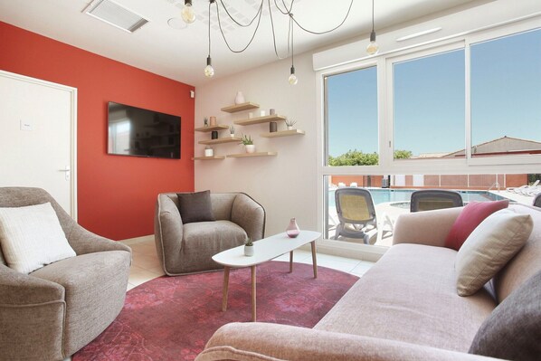 A modern living room with a flat-screen TV, comfortable seating, a coffee table, and a view of an outdoor swimming pool.