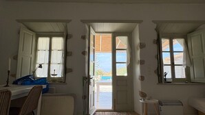 Una habitación con una puerta blanca que da a un balcón, una mesa pequeña con una lámpara, una silla de mimbre y un sofá blanco.