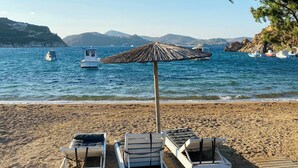 Una playa con sillas de descanso y una sombrilla de paja, con vistas al mar tranquilo y colinas lejanas.