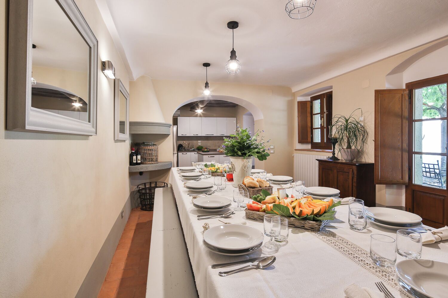 Une longue table à manger, dressée avec des assiettes, des verres et une variété de plats, est bordée de meubles de cuisine et d’une fenêtre offrant une vue agréable.