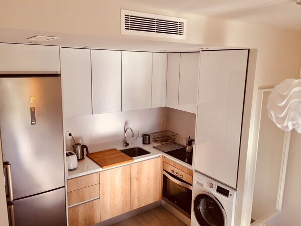 A compact kitchen with a stainless steel refrigerator, a built-in oven, a washing machine, and wooden cabinets.