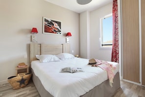 A neatly made bed with a book and a hat on it, a small bedside table with a lamp, and a window with curtains. - Maisonette in Aigues-Mortes near Camargue (Aïgues Mortes)