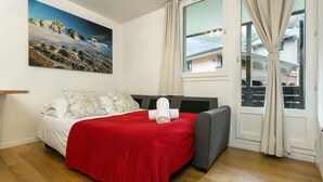 Un dormitorio con una cama, una manta roja, un sillón gris, una ventana con cortinas y un cuadro en la pared.