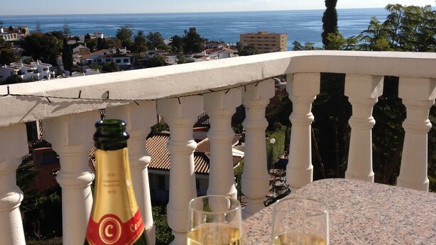 Eine Flasche Cuvée EXTRA Codorniu Wein auf einem Balkon mit zwei Weingläsern, mit Blick auf die Küste.