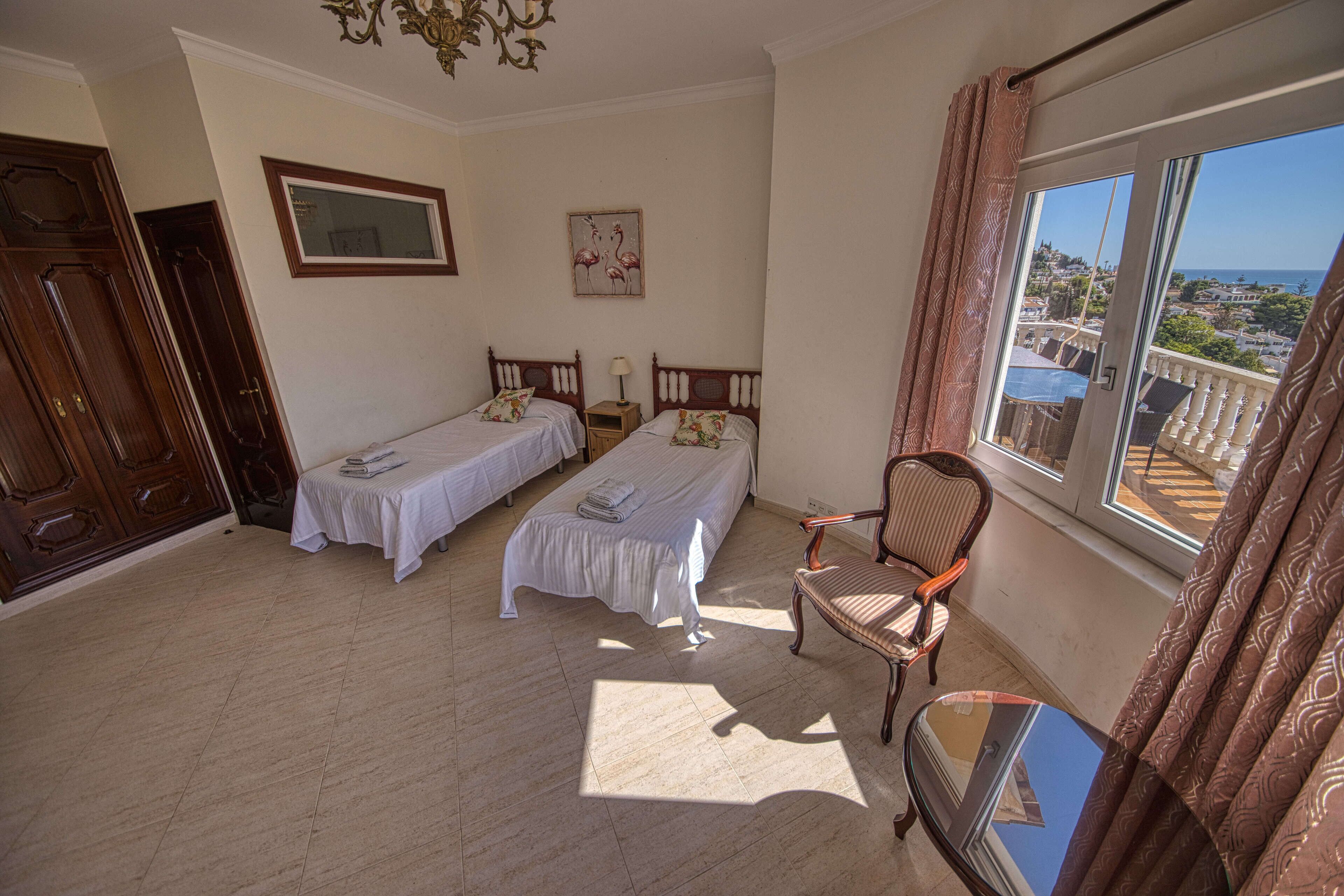 A room with two single beds, a wooden chair, a small table, and a window with a view of the sea.