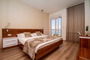 Une chambre à coucher avec un lit en bois, du linge de lit blanc, une table de chevet et une vue sur l’extérieur grâce à une grande fenêtre.