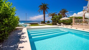 A swimming pool with clear blue water, surrounded by lounge chairs and umbrellas, with a view of the sea and palm trees in the background.
