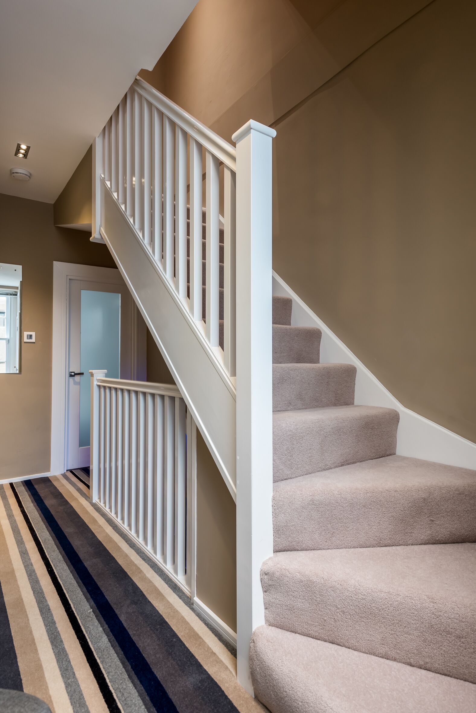 Una escalera con peldaños cubiertos de moqueta beige y barandillas blancas, que conduce a una puerta con un panel de vidrio satinado.