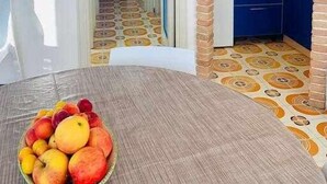 A room with a dining table, a fruit bowl, a kitchen area with blue cabinets, and a patterned floor. - Beachfront three room apartment with garden "Ostrica" (Alghero)