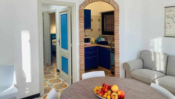 A dining area with a table set for two, a fruit bowl on the table, and a kitchen visible through an archway. - Beachfront three room apartment with garden "Ostrica" (Alghero)