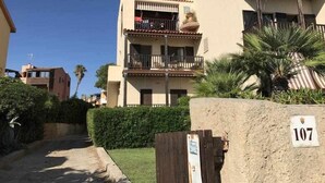 A multi-story residential building with balconies, a stone wall with a number plaque, and surrounding greenery. - Beachfront three room apartment with garden "Ostrica" (Alghero)
