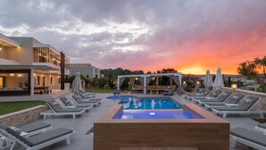 A modern outdoor pool area with lounge chairs and umbrellas, surrounded by a landscaped garden and a building with a terrace. -  Elaida Villa, Refined Group Escape • Pool & Play (Rethymno)