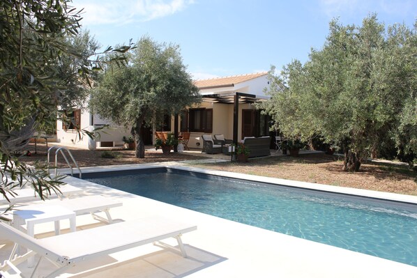 A swimming pool with lounge chairs, surrounded by olive trees and a house with outdoor furniture.