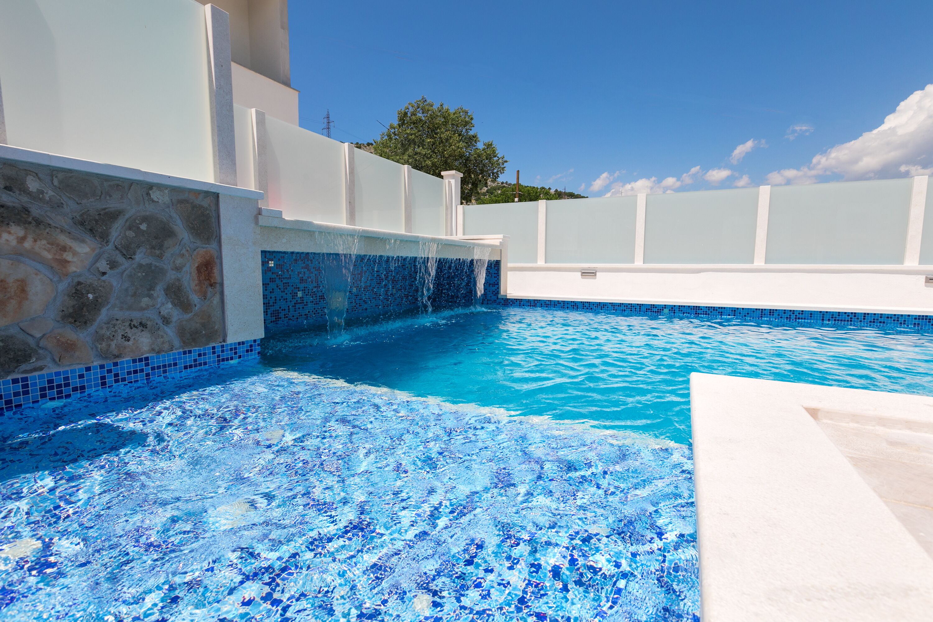 Una piscina con agua azul cristalina, un muro de piedra y un borde de azulejos.