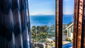 Vista d'una finestra cap a un paisatge costaner amb cel serè, una muntanya allunyada i un edifici amb balcó.