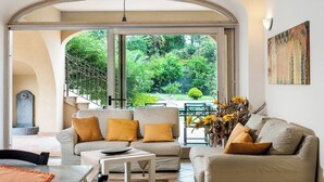 A living room with a sofa and armchairs, a coffee table, and a view of a garden through large sliding glass doors. - Villa Mareneve, Villa with Private Pool, Etna Sea view. CIN EN087048C2PTJBZY2J (Santa Venerina, Catania)