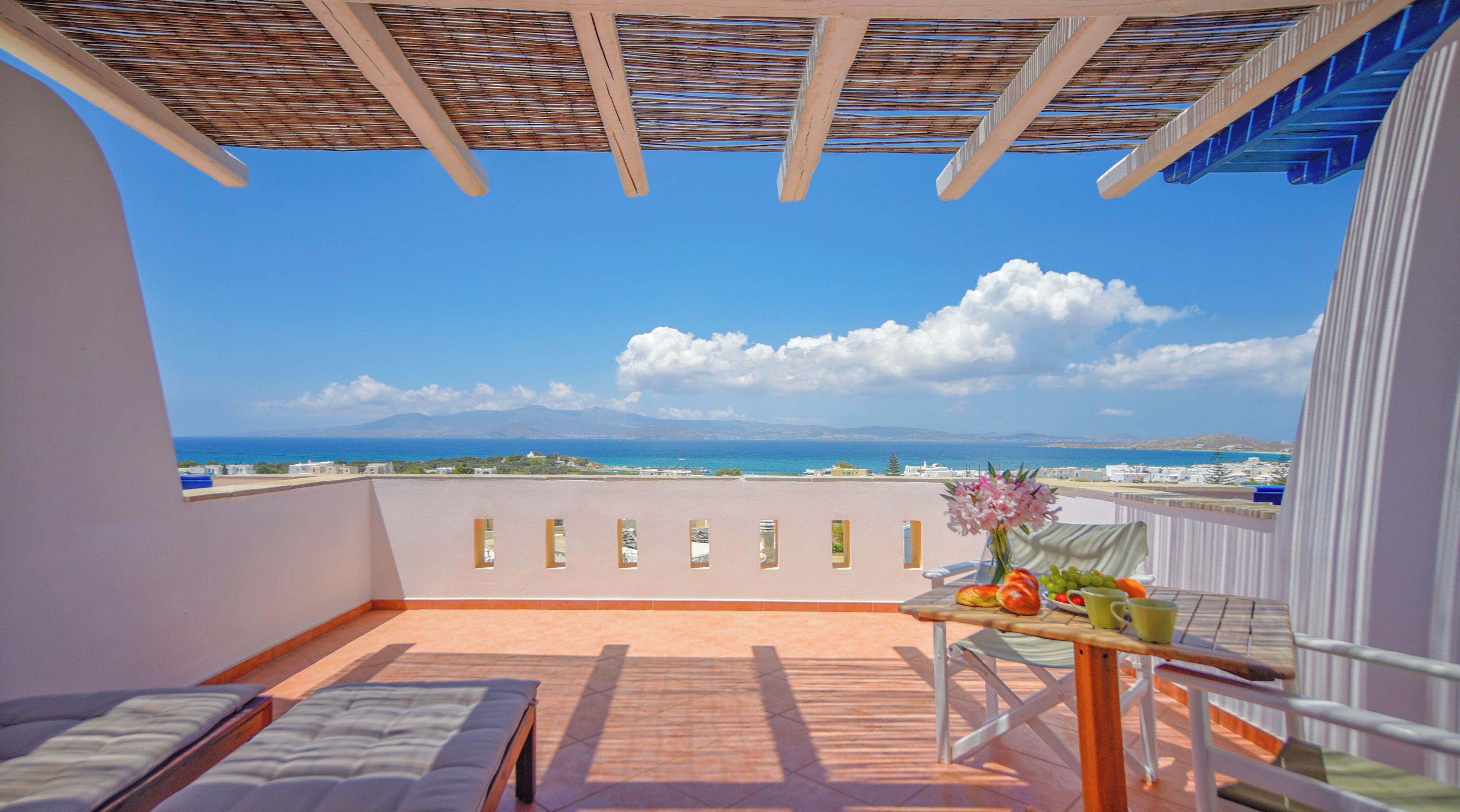 A balcony with a view of the sea, a table with fruits and a vase, and a bed.