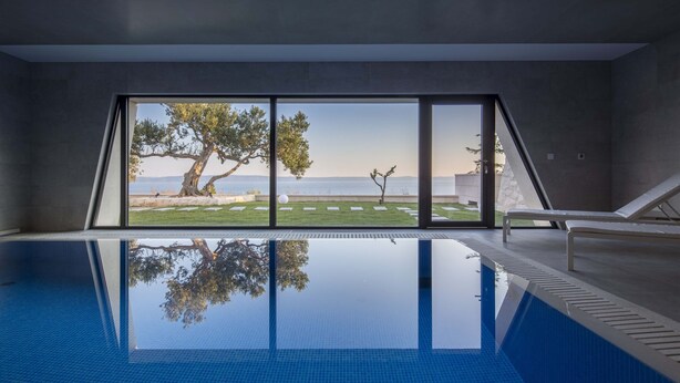 A modern indoor swimming pool with clear glass walls, offering a view of a landscaped area with trees and a distant body of water. - 5*Familiy Villa,Seaview,Indoor&Outdoor Heated Pools (Makarska)