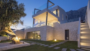 A modern two-story house with a stone facade, large glass windows, and a well-lit outdoor area with a tree and steps leading to the entrance. - 5*Familiy Villa,Seaview,Indoor&Outdoor Heated Pools (Makarska)