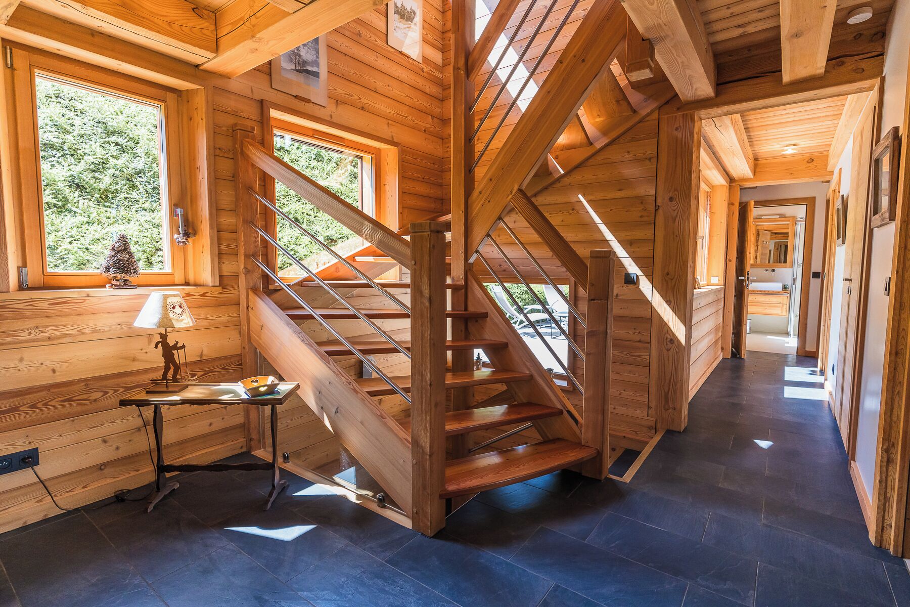 A wooden interior with a staircase leading to an upper level, a small table with a lamp, and a view of the outdoors through large windows.