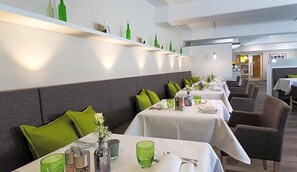 A restaurant with multiple tables, each set with green glassware, white tablecloths, and grey upholstered chairs.