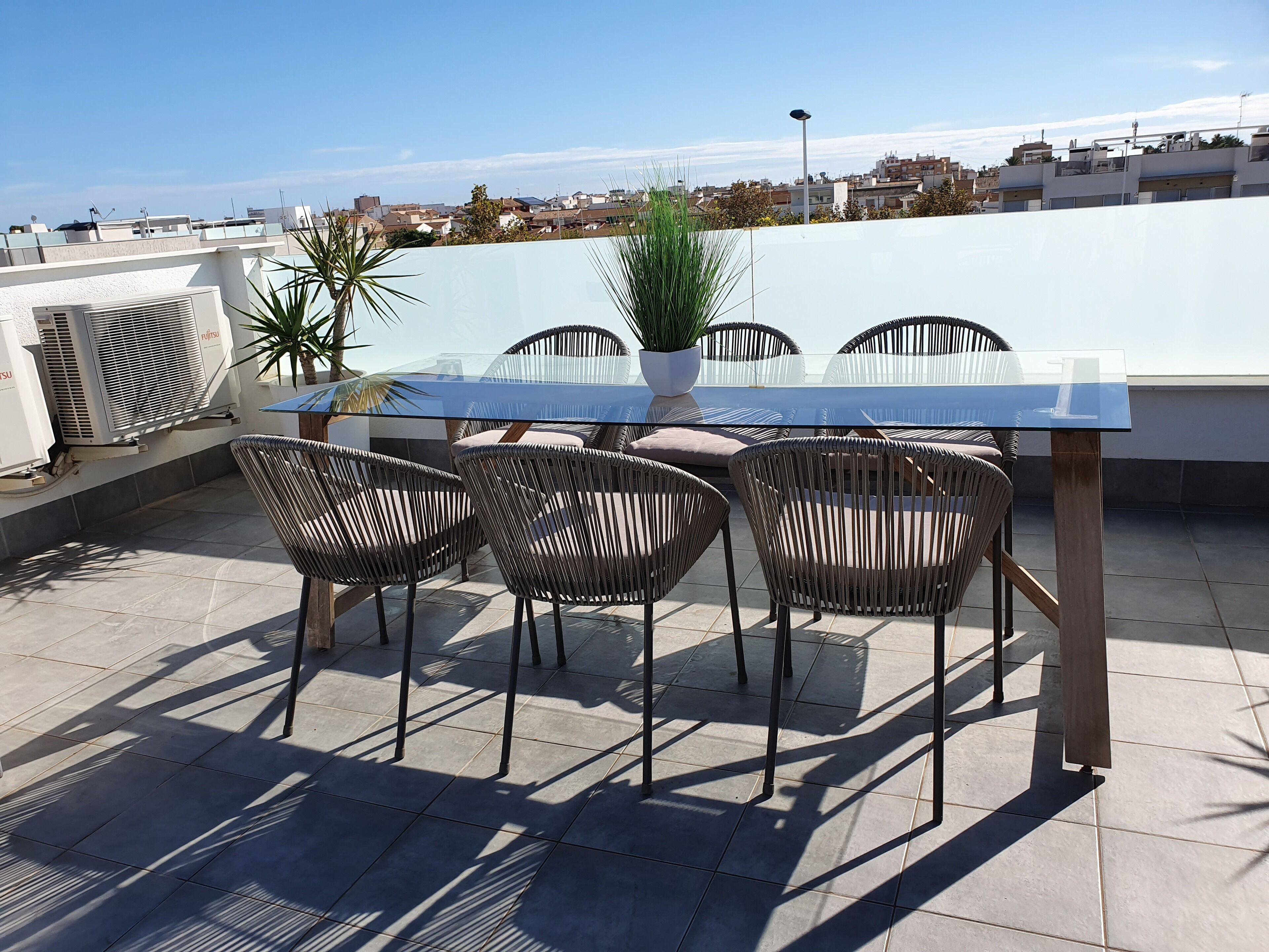Une terrasse sur le toit, dotée d’une table en verre et de chaises en osier, entourée de plantes en pot et de climatiseurs.