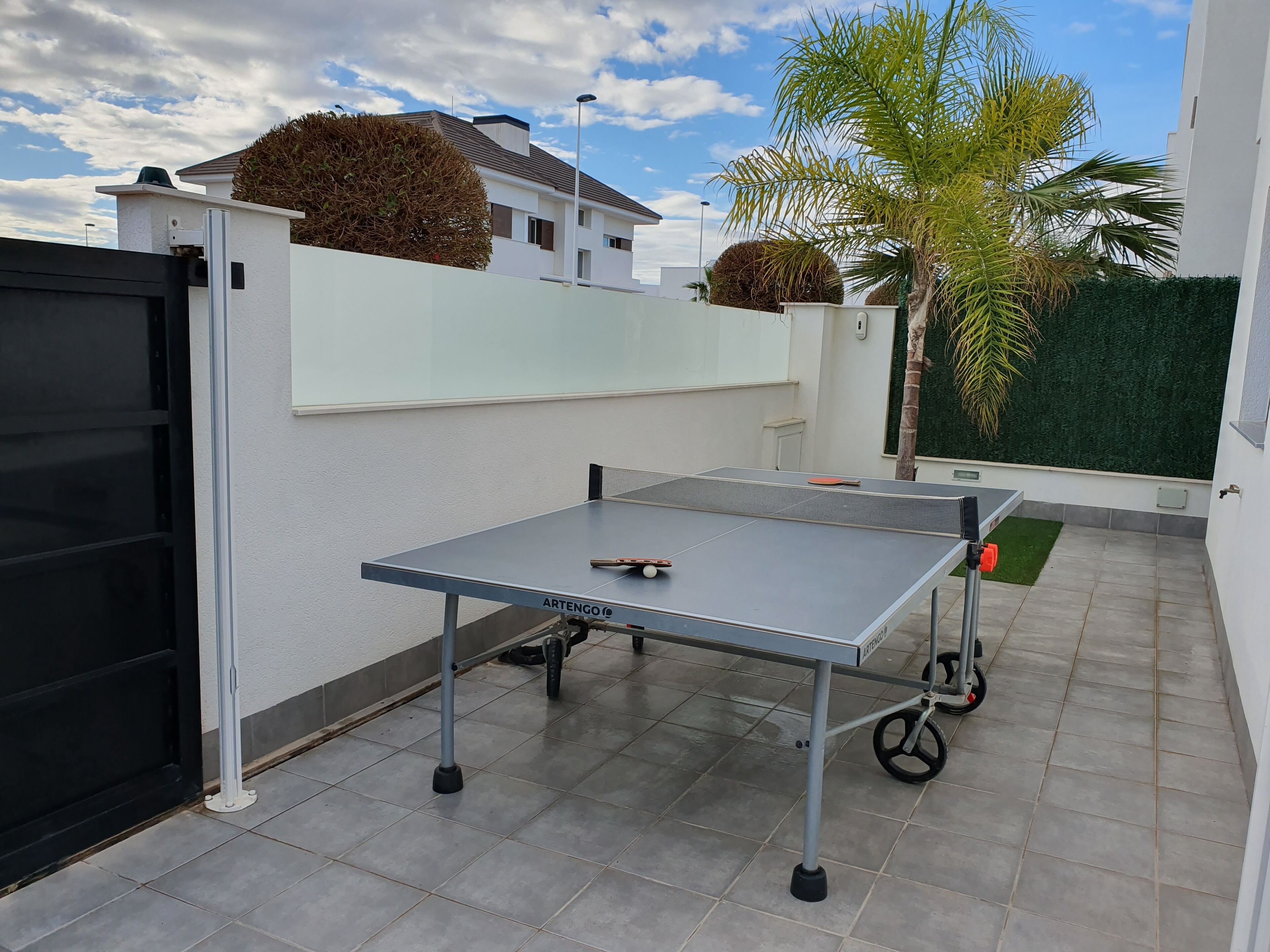 An outdoor ping pong table with wheels, a black gate, and a white wall with a green hedge.