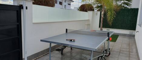 An outdoor ping pong table with wheels, a black gate, and a white wall with a green hedge.