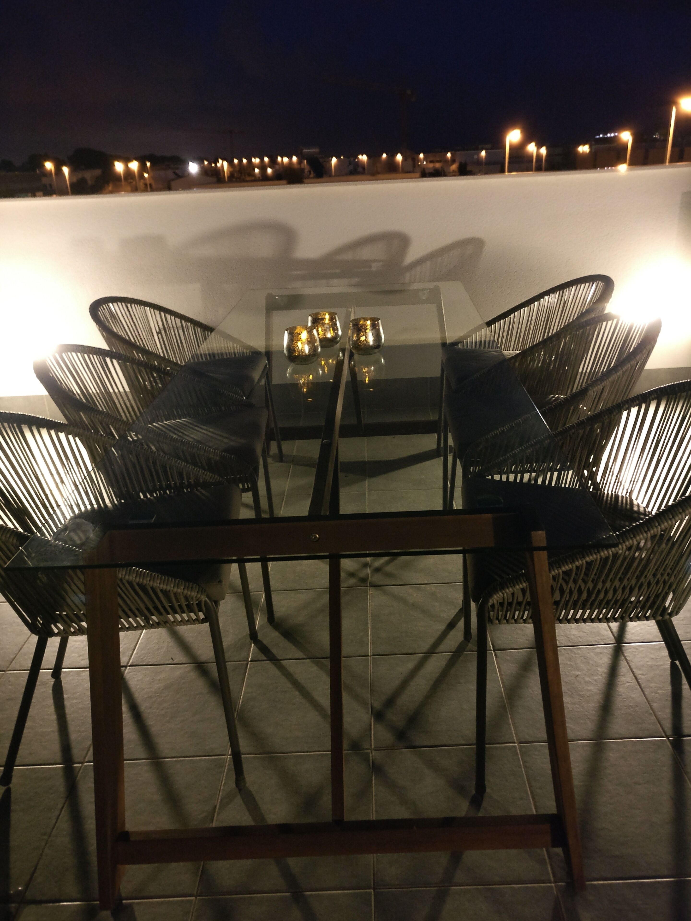 An outdoor dining area with a glass table and metal chairs, surrounded by a tiled floor and a low wall.