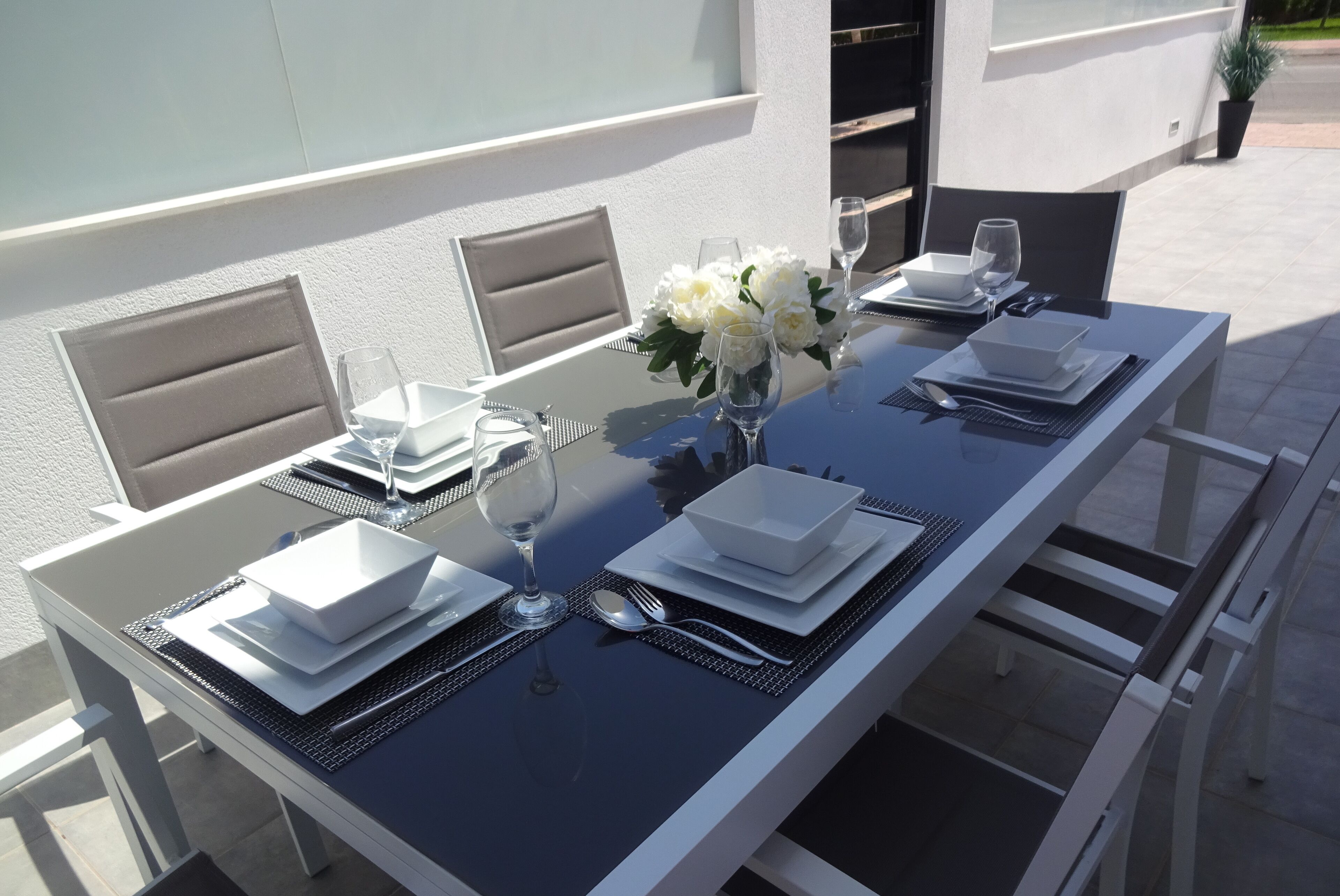 A dining table set with white plates, bowls, cutlery, and glasses, accompanied by a vase of white flowers.