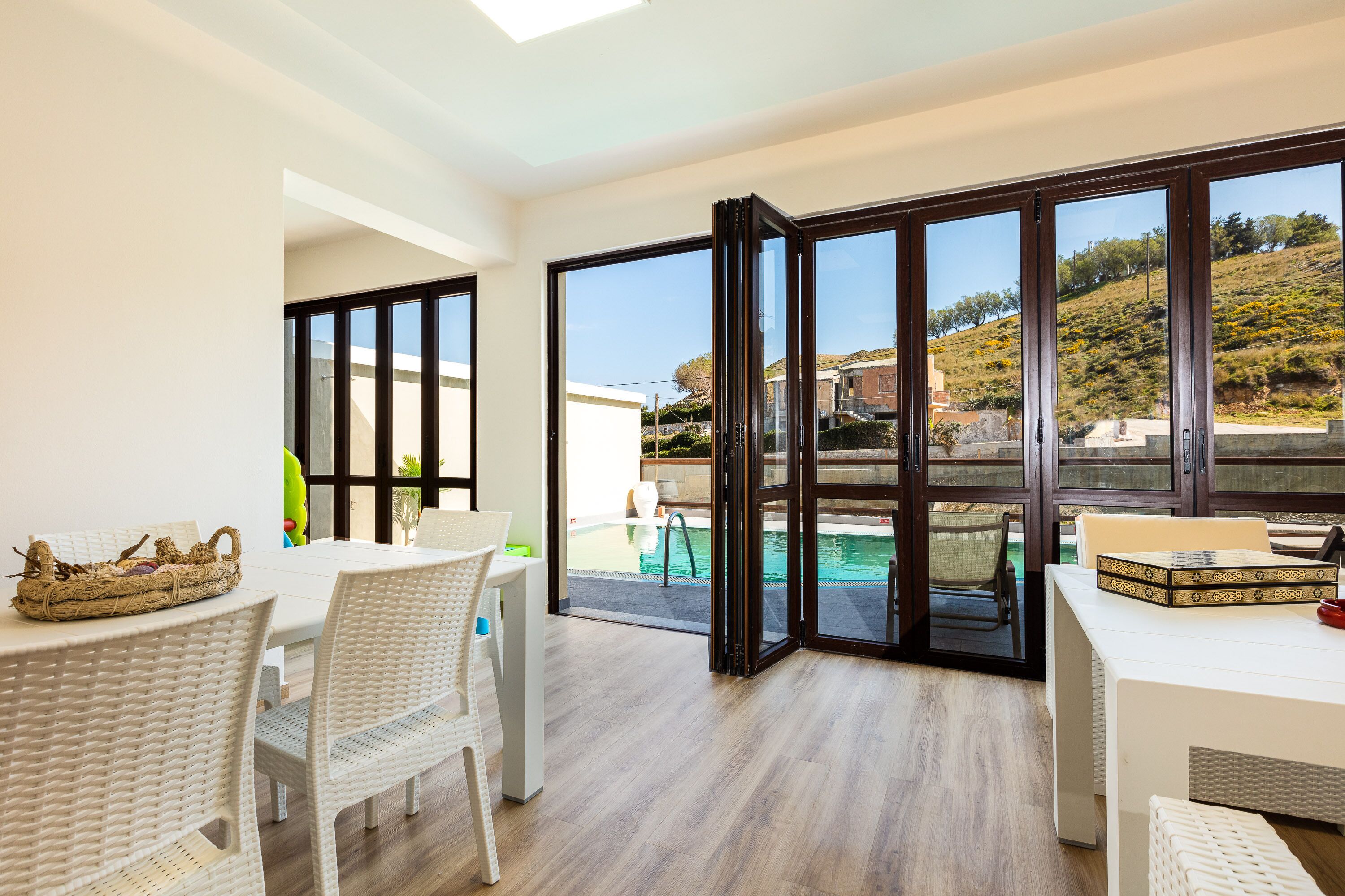 A modern dining area with a pool view, featuring a white table, wicker chairs, and a wicker basket.