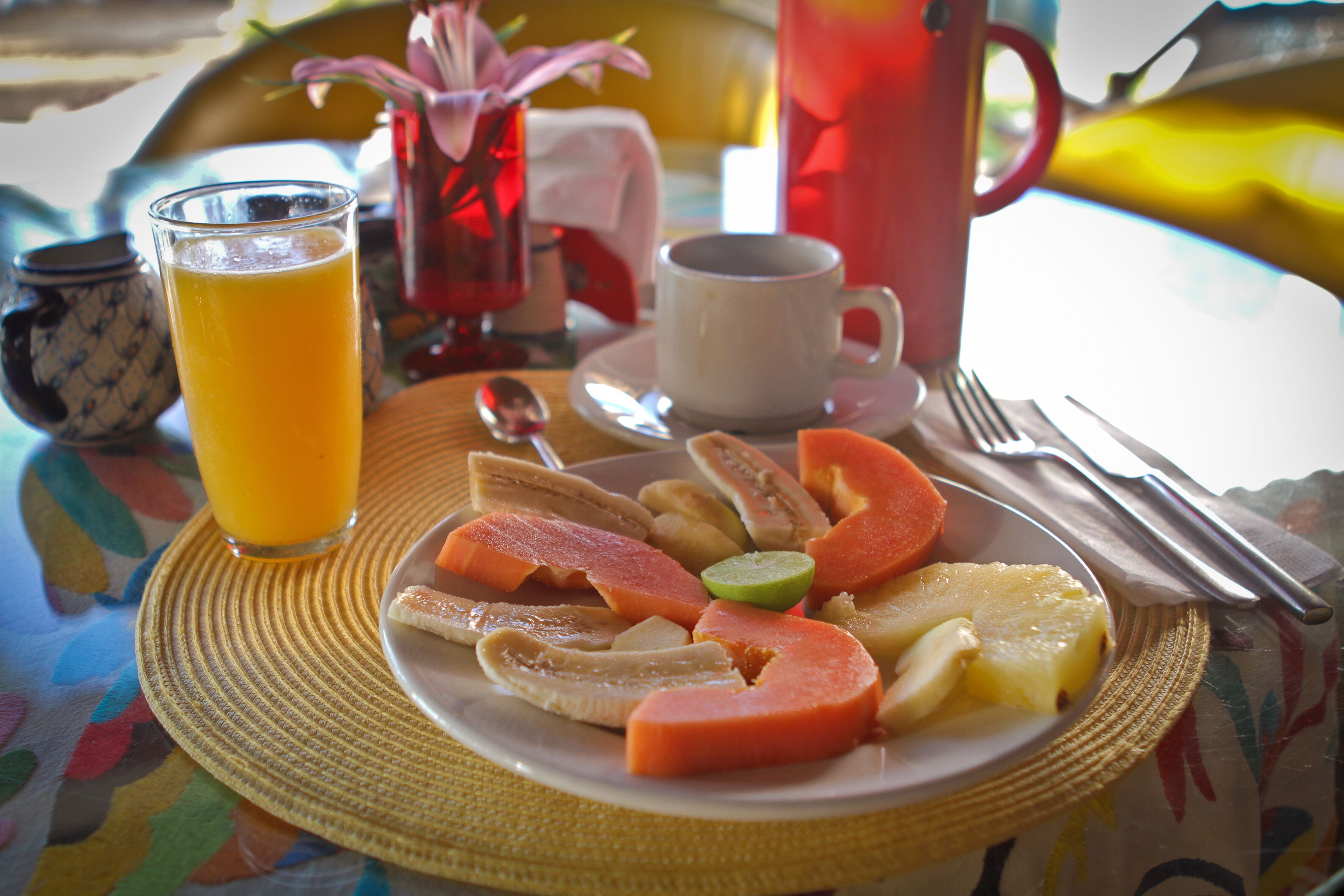 A breakfast setting with a plate of fruit, a glass of orange juice, a cup, and a vase with flowers.