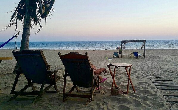 A beach with two wooden chairs and a small table, a palm tree, and a wooden structure in the background.