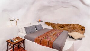A bedroom with a white bed, a patterned throw, a small wooden table with books, a wall-mounted lamp, and a textured wall. - Traditional Farmers Secret Cave #1 (SANTORINI)