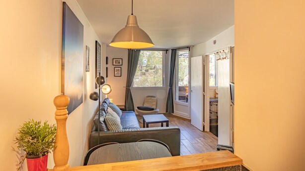 Un salon moderne avec un canapé, une table à manger et un luminaire suspendu. Un escalier mène à un autre niveau, et une porte se trouve sur la droite.
