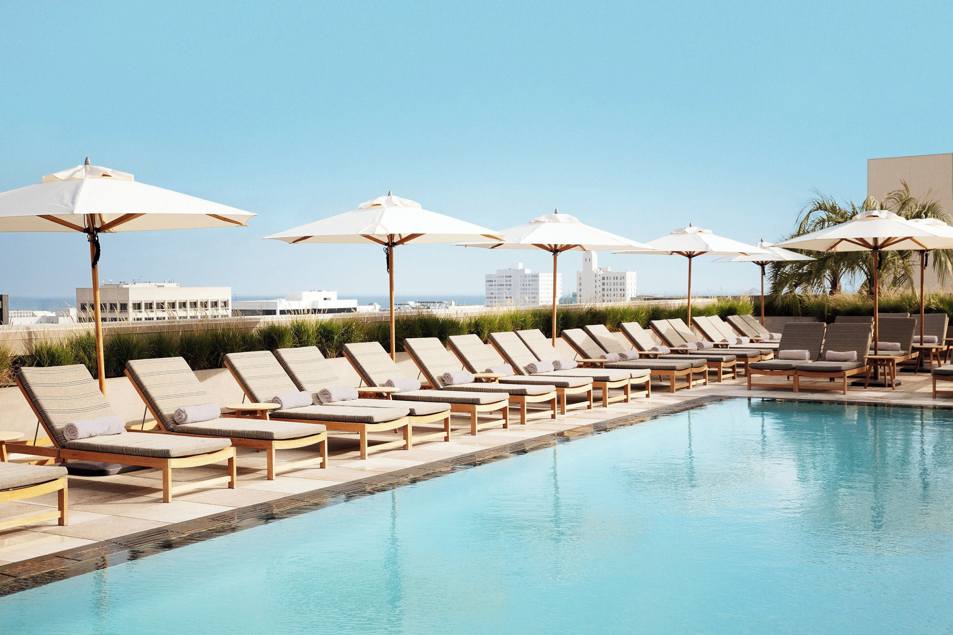 A poolside area with lounge chairs and umbrellas, a clear sky, and buildings in the background.
