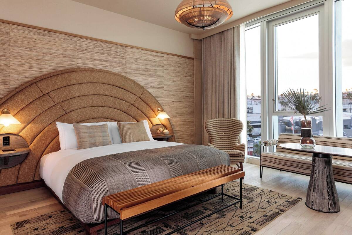 Una habitación de hotel moderna con una cama grande, un banco de madera, una mesa redonda y vistas de la ciudad a través de amplios ventanales.