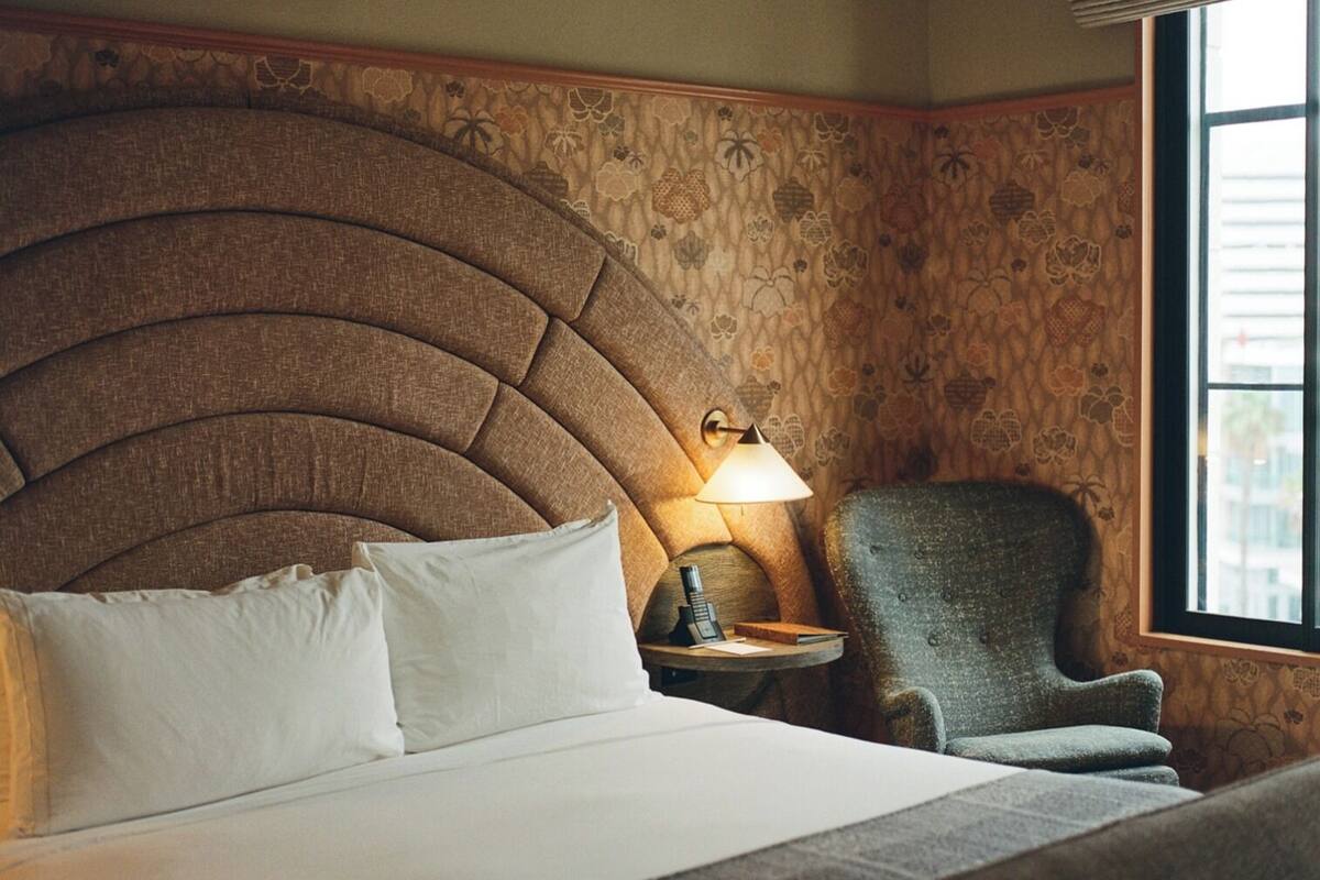Habitación de hotel con un cabecero grande y capitonado, una lámpara de noche, una silla y una ventana con vistas.