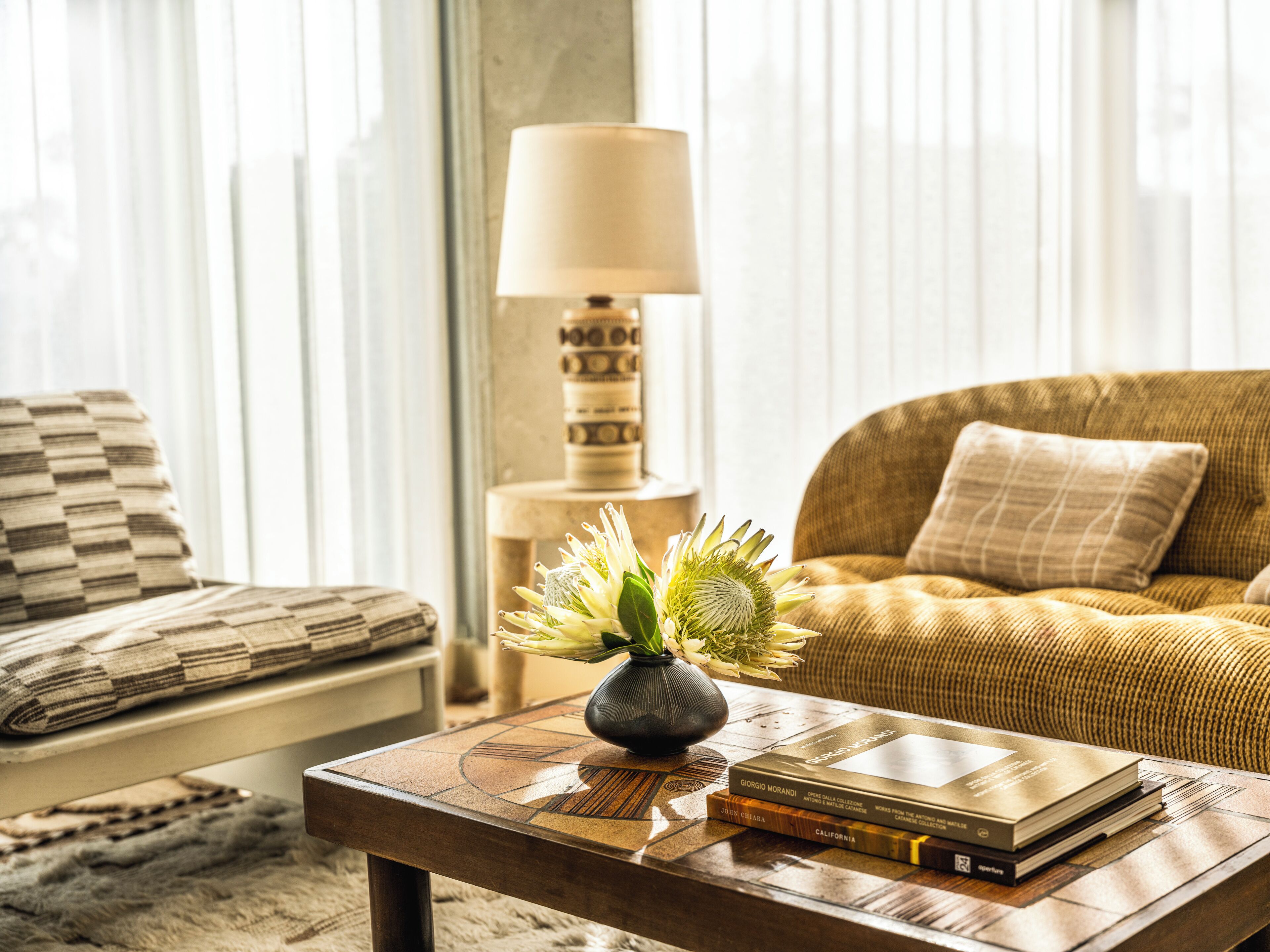 A living room with a sofa, a coffee table with books and a vase of flowers, and a lamp in the background.