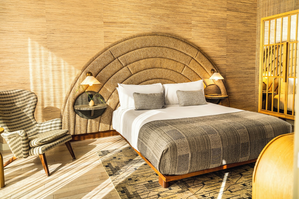 A bedroom with a curved headboard, a bed with white linens, two bedside lamps, a striped armchair, and a patterned rug.