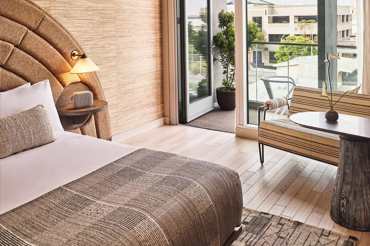 Un dormitorio moderno con una cama grande, una mesita de noche con lámpara, cabecera de madera y vistas a la ciudad a través de grandes ventanales.