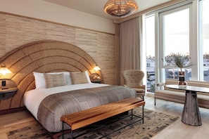 A modern hotel room with a large bed, a wooden bench, a round table, and a city view.