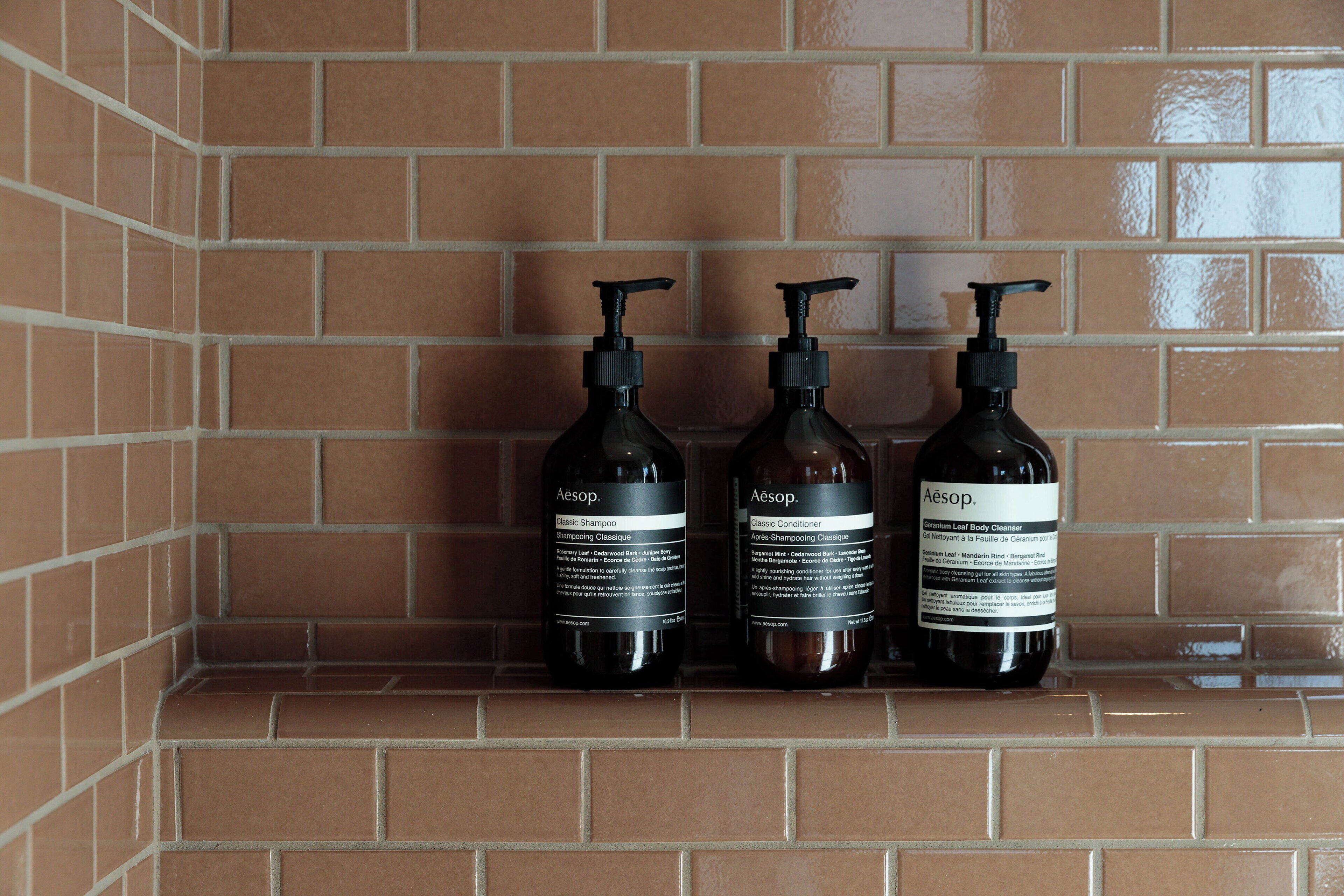 Three bottles of Aesop Classic Shampoo and Conditioner on a tiled shelf.